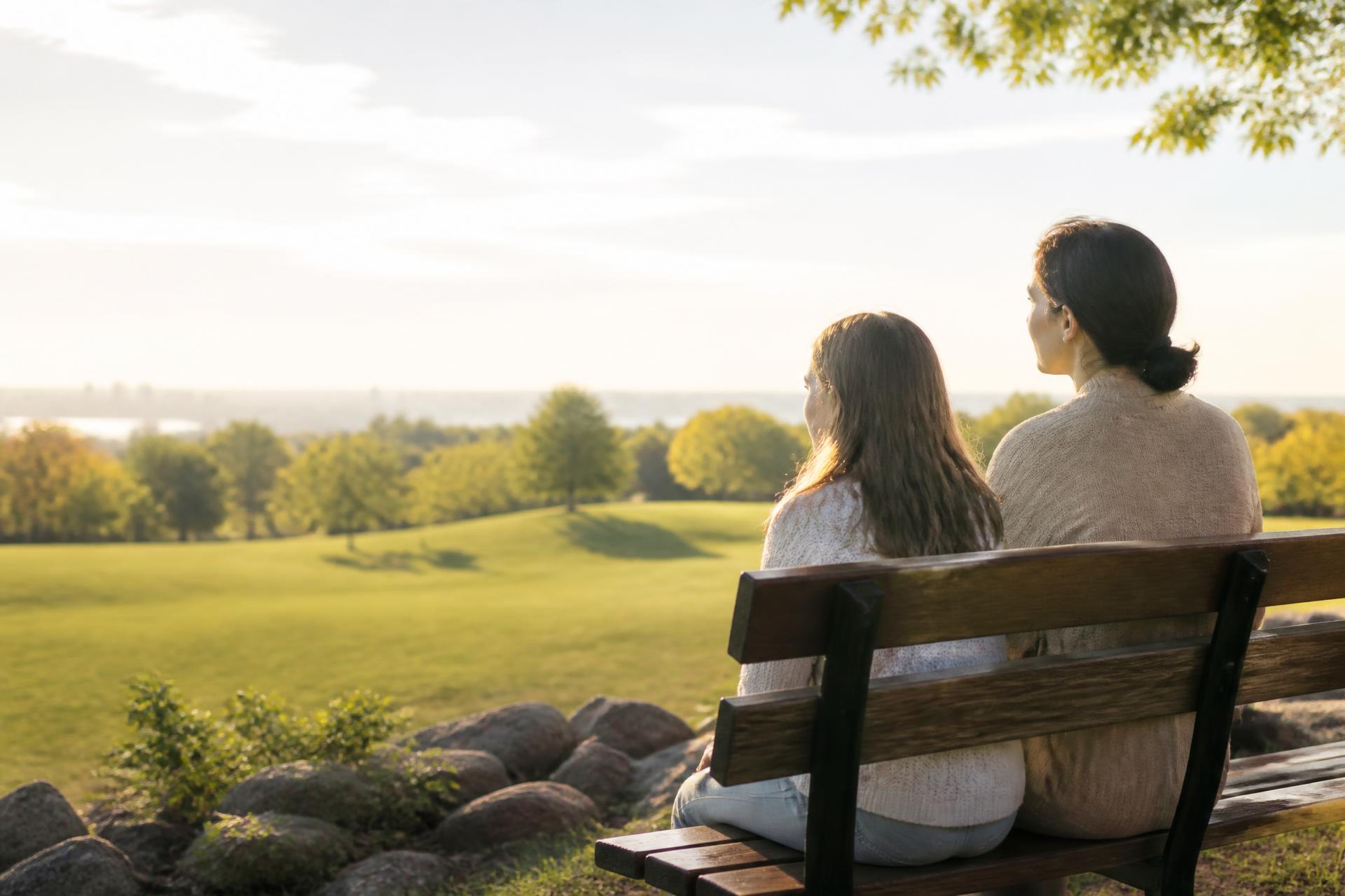 Two women sitting together on a park bench in soft morning light, one looking away and the other turned gently towards her.
